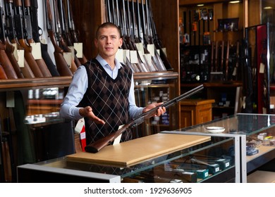 Portrait Of Gun Store Salesman Showing Collectible Old Rifled Musket On Background With Rack Full Of Vintage And Modern Firearms