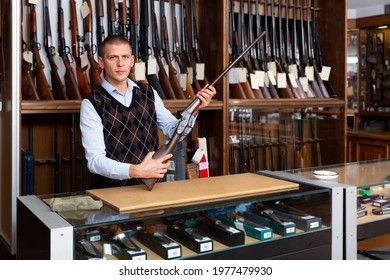 Portrait Of Gun Store Salesman Showing Collectible Old Rifled Musket On Background With Rack Full Of Vintage And Modern Firearms