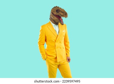 Portrait of guy in funky costume with dinosaur face posing in studio. Man in stylish bright yellow suit and funny ugly T Rex mask standing with hand in pocket on blue background. Crazy fashion concept