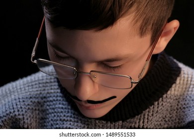 Portrait Of A Guy In Glasses With A Mustache Stuck On. A Teenager Lowered His Head While Reading A Book Or Other Mental Work. Black Background. Theatrical Image Of An Adult.