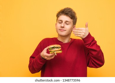 Portrait of a guy holding a burger in his hands and enjoying his eyes closed, isolated on a yellow background. Young man with fast food in his hands is on a yellow background in the studio