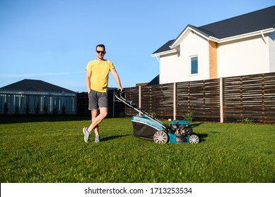 Portrait Of Guy With A Lawn Mower On A Back Yard Of Private House, He Is In Contemporary Sunglasses And In Yellow Casual T-shirt Looks At Camera And Smiles. Care About Backyard