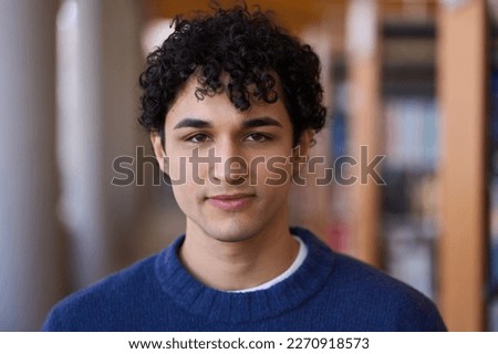 Portrait of handsome 20 years old young Latin American man, wearing blue knitted sweater, confidently looking at camera, standing against blurred background of bookshelves in a library or a bookstore