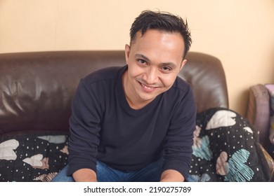 Portrait Of A Handsome 30 Year Old Asian Man Wearing Blue Top Smiling Vividly While Sitting On A Brown Couch In His Living Room.