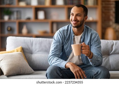 Portrait of handsome african american man resting on couch at home with cup of coffee, young black guy sitting on sofa in living room interior, looking away and enjoying hot drink, copy space - Powered by Shutterstock