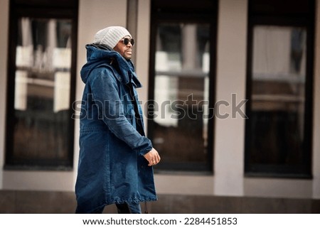 Portrait of handsome African American man walking in city, wearing stylish outfit parka coat, knitted hat and sunglasses. Street style fashion male model