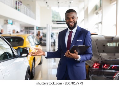 Portrait Of Handsome African American Salesman At Workplace In Car Showroom, Happy Smiling Black Auto Dealership Manager In Suit With Clipboard In Hands Demonstrating New Vehicles, Copy Space