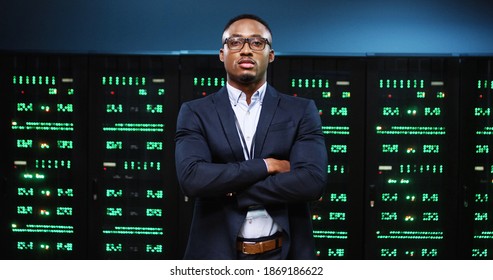 Portrait Of Handsome African American Serious Male IT Security Engineer Standing In Big Data Center With Server Racks And Looking At Camera. Man Analytic In Glasses At Work In Server Room Tech Concept