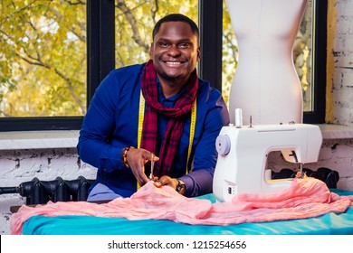 portrait of a handsome african man smiling seamstress with sewing machine.Afrio American man stylish designer working in tailor workshop mannequin,table measuring tape in room against autumn window