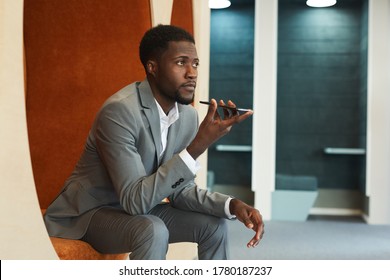 Portrait of handsome African-American businessman recording voice message via smartphone while sitting in lounge zone of modern office or coworking space - Powered by Shutterstock