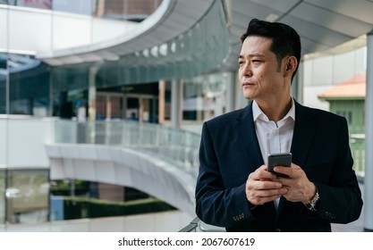 Portrait Of Handsome Asian Businessman Using Mobile Phone Outside Office. 

Serious Business Man In Blue Suit Standing At Modern Balcony And Typing Text Message On His Smartphone While Looking Away.