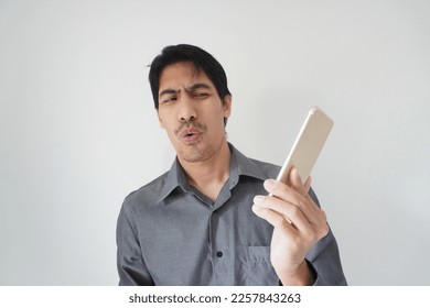 Portrait of a handsome Asian male looking at his smartphone screen with a funny, tired facial expression, isolated background. Concept of businessperson in frustration stressed, bored with overwork. - Shutterstock ID 2257843263