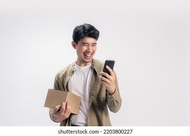 Portrait Handsome Asian Man In Casual Outfit, With Holding A Box And Happy Using Online Shopping On Smartphone, With An Exciting Smile On Isolated White Background.