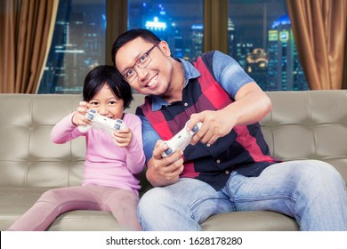 Portrait Of Handsome Asian Man Playing Video Games Cheerfully & Enthusiastically With His Daughter In Their Family Room At Night