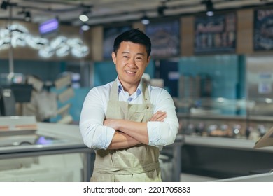 Portrait Of Handsome Asian Man Salesman In Apron Standing And Looking At Camera In Grocery Store Supermarket. Male Worker Seller Or Small Business Owner. Indoor. Food, Groceries Market Cheerful Smile.