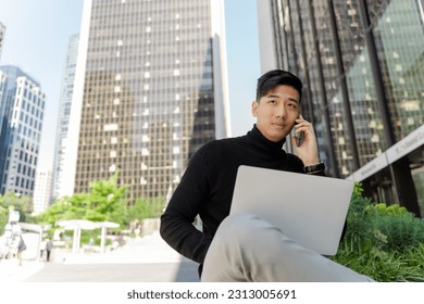 Portrait of handsome asian man using laptop, talking on mobile phone, outdoors. Young chinese student preparing for exam, sitting on city street - Powered by Shutterstock