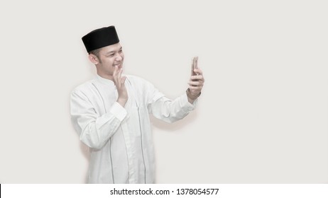 Portrait Of Handsome Asian Muslim Man With Head Cap And Using Smartphone Taking A Video Call And Waving Smile At Phone -image