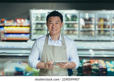 Portrait Of Handsome Asian Staff Man Salesman In Apron Standing And Looking At Camera In Grocery Store Supermarket. Male Worker Seller Or Small Business Owner. Indoor. Food, Groceries Market Smile