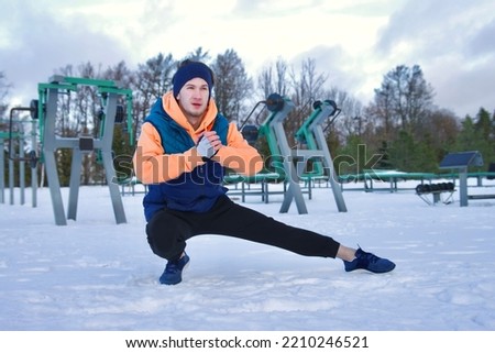 Portrait of handsome athletic fit fitness guy, young man is stretching his body, muscles before workout, training outdoors at gym at winter cold snow day, snowy frosty weather, warming up.