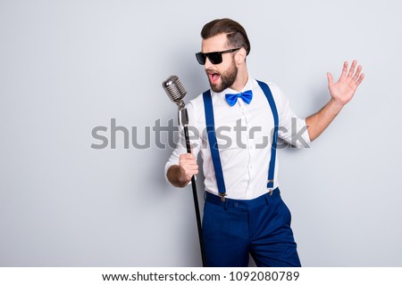 Portrait of handsome attractive singer in blue pants with suspenders and black glasses, singing hit with open mouth in microphone gesture with hand isolated on grey background