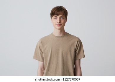 Portrait Of Handsome Attractive Young Man With Short Haircut And Blue Eyes Wears Beige T Shirt Standing And Smiling Isolated Over White Background Looks Directly In Camera