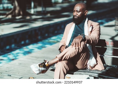 A Portrait Of A Handsome Bald Bearded Black Senior Man In A Fancy Pinkish Tailored Suit Sitting On A Wooden Street Bench In A Park And Thoughtfully Looking Away; With A Copy Space Place On The Left