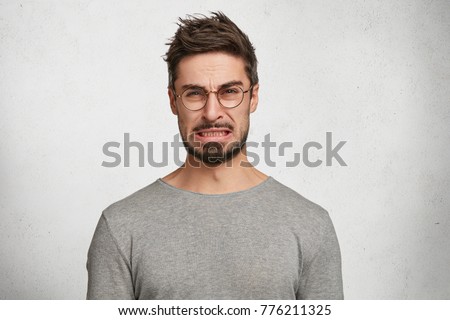 Portrait of handsome bearded male with disgusting expression sees sour lemon in front, frowns in displeasure, isolated over white concrete background. Male expresses aversion towards something