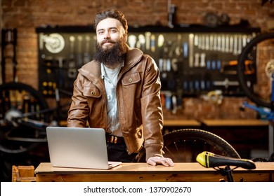 Portrait Of A Handsome Bearded Man As Bicycle Store Owner Or Manager Standing With Laptop At The Bicycle Workshop