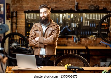 Portrait Of A Handsome Bearded Man As Bicycle Store Owner Or Manager Standing With Laptop At The Bicycle Workshop