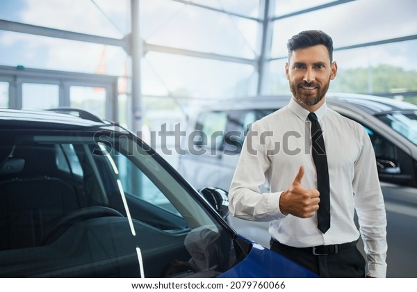 Portrait of handsome bearded\
man in formal clothes showing thumb up and smiling on camera while\
standing at auto salon. Male customer making expensive\
purchase.