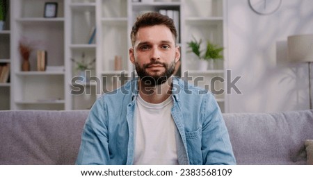 Portrait of handsome bearded man in headphones sitting on couch posing and looking at the camera, indoors