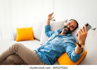 Portrait Handsome Bearded Man Wearing Glasses,headphones Listening To Music At Modern Home. Guy Sitting At Sofa, Holding Smartphone And Relaxing. Happy Man In Earphones With Smartphone