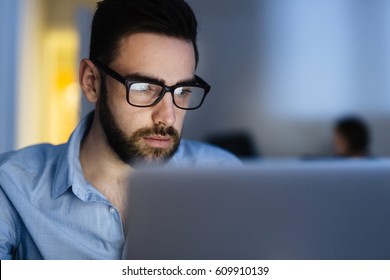 Portrait Of Handsome Bearded Man Wearing Glasses Working With Laptop In Dark Office Late At Night, His Face Lit Up By Screen