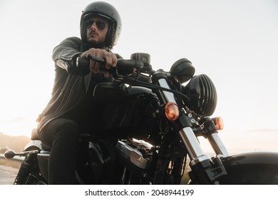 Portrait Of Handsome Biker Sitting On His Vintage Motor Bike, Wearing Leather Jacket, Helmet And Sunglasses