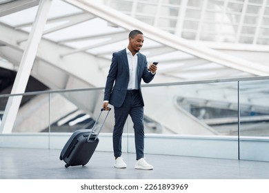 Portrait Of Handsome Black Businessman Walking With Suitcase In Airport And Using Smartphone, Young African American Man In Suit Browsing Internet On Cellphone While Going To Flight Gate, Copy Space