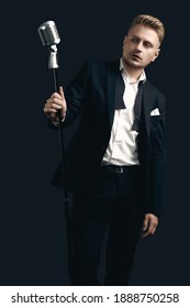 Portrait of handsome blond man singer in elegant tuxedo and bow tie posing with vintage microphone on black studio background