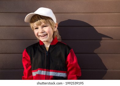 Portrait of a handsome boy 5 years old in a red uniform of a mechanic and a white cap against the background of a garage door. smiles with a happy face. Light and shade. Little helper. Be like dad