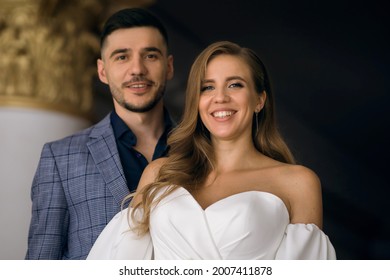 Portrait Of A Handsome Brunette Man With A Beard And A Young Woman With An Elegant Hairstyle In A White Dress. A Loving Couple Is Happy And Smiling. Heterosexual Couple Indoors