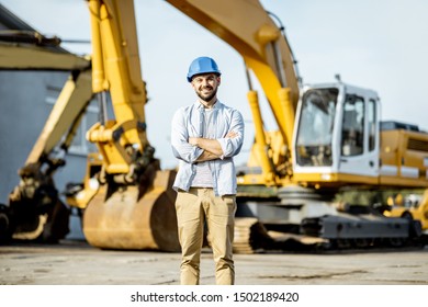 Portrait Of A Handsome Builder Standing On The Open Ground Of The Shop With Heavy Machinery For Construction