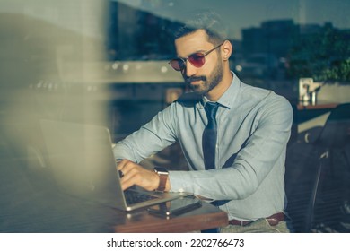 Portrait Of Handsome Business Man Wearing Sunglasses Working On Laptop In Modern Office. Through Glass View.