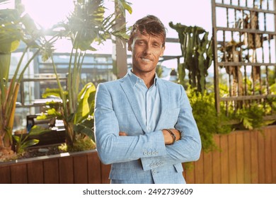 Portrait of a handsome businessman with suit, poses with arms crossed outdoors near plants, broadly smiles looking at camera. - Powered by Shutterstock