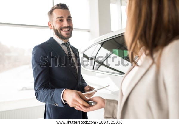 Portrait of handsome car salesman giving car\
keys to young woman standing next to white shiny luxury car in\
dealership showroom