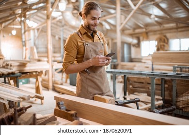 Porträt eines gut aussehenden Zimmermanns, der mit einem Smartphone steht und während der Holzarbeiten in der Werkstatt bremst