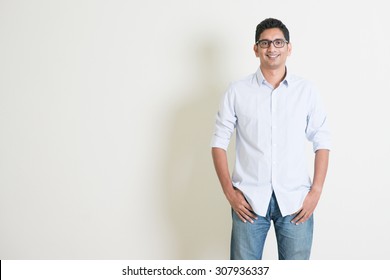 Portrait Of Handsome Casual Business Indian Guy Smiling, Hands In Pocket, Standing On Plain Background With Shadow, Copy Space At Side.