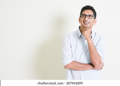 Portrait Of Handsome Casual Business Indian Man Smiling And Thinking, Eyes Looking Upwards, Standing On Plain Background With Shadow, Copy Space At Side.