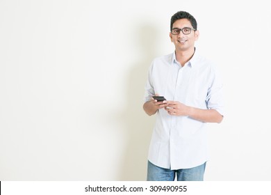 Portrait Of Handsome Casual Business Indian Man Using Smartphone, Social Media Concept, Standing On Plain Background With Shadow, Copy Space At Side.