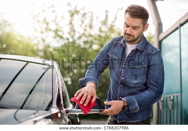 Portrait of handsome Caucasian bearded man in\
jeans shirt, wiping side mirror with red microfiber rag at outdoor\
car wash self station.