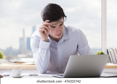 Portrait of handsome caucasian guy using notebook computer at office desk with coffee cup, paperwork and other items. Blurry city background - Shutterstock ID 476325742
