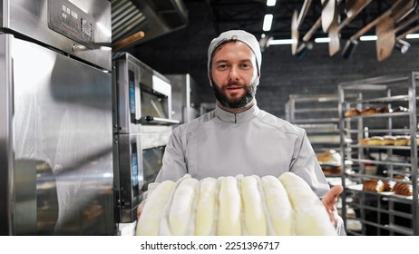 Portrait of handsome Caucasian male in apron and hat standing in front of camera, holding tray with sweet bakery and smiling in kitchen of bakehouse. Man worker of bakeshop. Confectionery concept. - Shutterstock ID 2251396717
