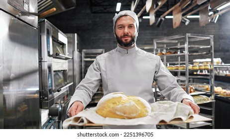 Portrait of handsome Caucasian male in white apron and hat standing in front of camera, holding tray with fresh just-baked bread and smiling in kitchen of bakehouse. Man in bakeshop showing baking.